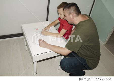 A child returns from school wearing red T shirt and sits down white table to do their schoolwork and homework with help of their father. A child returns from school wearing red T shirt and sits down white table to do their schoolwork and homework with help of their father. 143777788