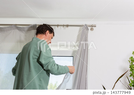 A man is hanging a curtain on the rod after washing it. It is spring, and he is doing household chores in a room with a window and some greenery A man is hanging a curtain on the rod after washing it. It is spring, and he is doing household chores in a room with a window and some greenery 143777949