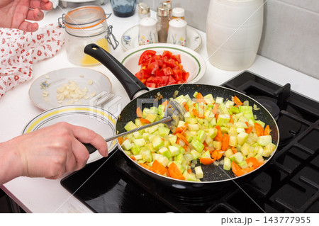 Hands stir chopped squash, carrots, and tomatoes in a pan on a kitchen stove during preparation of squash caviar at home Hands stir chopped squash, carrots, and tomatoes in a pan on a kitchen stove during preparation of squash caviar at home 143777955