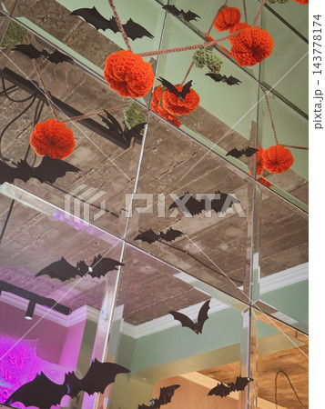 Halloween pumpkin decorations made from orange crochet yarn hang alongside black paper bats against mirrored wall. Pumpkin garland creates seasonal Background for festive design, craft projects 143778174