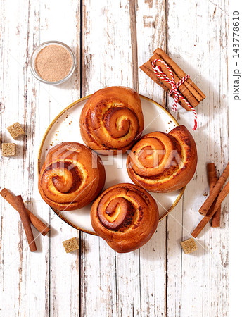Delicious hot buns rolls on a wooden table, accompanied by cinnamon sticks and burnt sugar, traditional autumn and winter dessert for the cold season. Ideal as a design template Delicious hot buns rolls on a wooden table, accompanied by cinnamon sticks and burnt sugar, traditional autumn and winter dessert for the cold season. Ideal as a design template 143778610