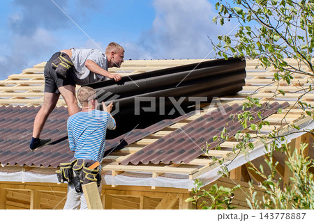 Roof installation and repair at heights with two workers fitting asphalt roofing panels over exposed framing on sloping wooden roof. 143778887