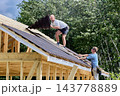 Roofing work at heights during installation of corrugated bitumen sheets on wooden battens above timber frame building under construction. 143778889
