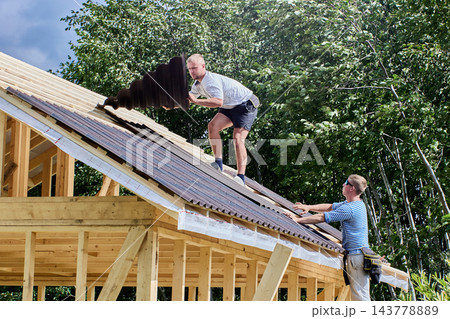 Roofing work at heights during installation of corrugated bitumen sheets on wooden battens above timber frame building under construction. 143778889