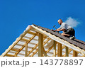 High-altitude work on timber roof with roofer using hammer to secure corrugated bitumen sheets above exposed wooden rafters. 143778897