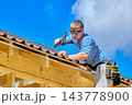Fixing corrugated asphalt roofing sheets with hammer and nails, construction worker secures roofing along timber eaves. 143778900