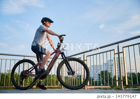 Man riding electric mountain bike on city promenade, side view cyclist against urban skyline and blue sky, active lifestyle and eco transport concept 143779147