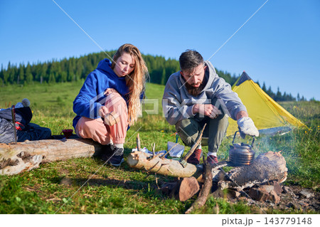 Man and woman by campfire in mountain meadow. Female tourist holding cup while husband tending to kettle on fire. Yellow tent and lush green hills in background, all under clear blue sky. Man and woman by campfire in mountain meadow. Female tourist holding cup while husband tending to kettle on fire. Yellow tent and lush green hills in background, all under clear blue sky. 143779148
