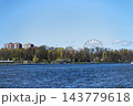Panoramic waterfront of upper pond in Kaliningrad with calm water, tree-lined shore, autumn foliage and distant Ferris wheel under bright clear blue daytime sky, Russia 143779618