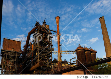 High angle telephoto landscape of historic blast furnace complex with orange conical exhaust stack cowper stoves brick smokestack and blue sky in Duisburg Ruhr region North Rhine Westphalia Germany High angle telephoto landscape of historic blast furnace complex with orange conical exhaust stack cowper stoves brick smokestack and blue sky in Duisburg Ruhr region North Rhine Westphalia Germany 143780266