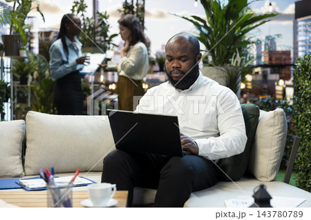 Businessman works on laptop in rooftop garden during networking event. African american man completes tasks on notebook in coworking space on outdoor terrace above business district Businessman works on laptop in rooftop garden during networking event. African american man completes tasks on notebook in coworking space on outdoor terrace above business district 143780789