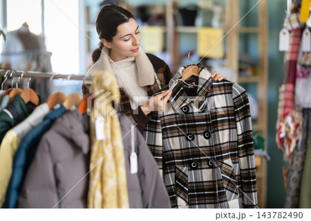 Interested happy young girl standing in clothing shop and looking new coat or jacket 143782490