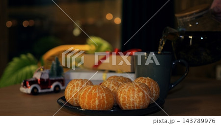 Steaming tea fills ceramic mug next to plate of peeled citrus fruits during festive evening. Wrapped presents with ribbons and toy car ornament establish warm cozy christmas ambiance at home. 143789976
