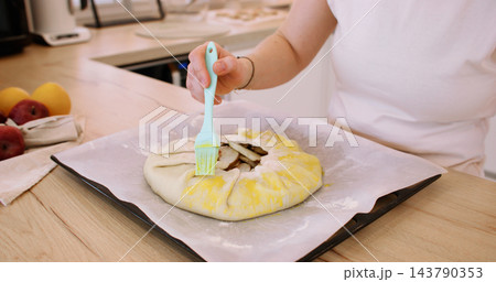 Sweet tart. Glazing pie crust. Woman baker hand brushing round galette with liquid yellow egg wash on baking sheet at home kitchen. Sweet tart. Glazing pie crust. Woman baker hand brushing round galette with liquid yellow egg wash on baking sheet at home kitchen. 143790353