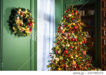 Christmas decoration idea. House decorated for holidays. Christmas wreath garland on green wall and Christmas tree with golden red ornaments decoration toys and garland lights in living room Christmas decoration idea. House decorated for holidays. Christmas wreath garland on green wall and Christmas tree with golden red ornaments decoration toys and garland lights in living room 143790373