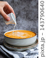 Bowl of creamy pumpkin soup topped with pumpkin seeds human hand pours fresh cream into the soup. Rich orange puree is served in rustic bowl and decorated with crunchy seeds creating an appetizing 143790640