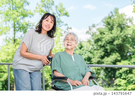 緑豊かな屋外で車椅子に乗る高齢の母親と付き添う娘の親子。 緑豊かな屋外で車椅子に乗る高齢の母親と付き添う娘の親子。 143791647