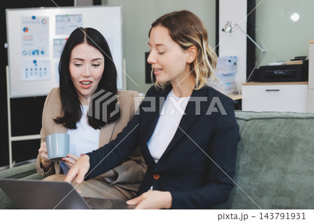 Two smiling women are comfortably sitting on a couch, sharing a moment and collaborating on a tablet, working together in a home office environment 143791931