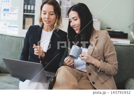 Two smiling women are comfortably sitting on a couch, sharing a moment and collaborating on a tablet, working together in a home office environment Two smiling women are comfortably sitting on a couch, sharing a moment and collaborating on a tablet, working together in a home office environment 143791932