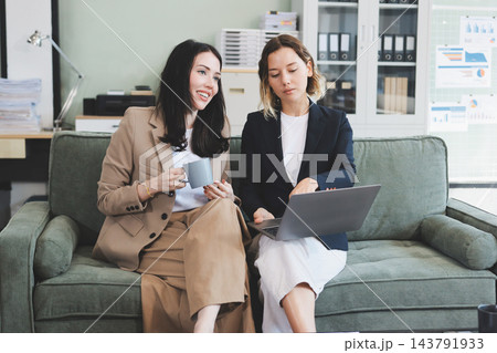 Two smiling women are comfortably sitting on a couch, sharing a moment and collaborating on a tablet, working together in a home office environment Two smiling women are comfortably sitting on a couch, sharing a moment and collaborating on a tablet, working together in a home office environment 143791933