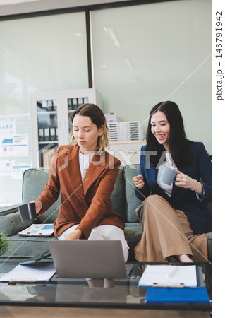 Two smiling women are comfortably sitting on a couch, sharing a moment and collaborating on a tablet, working together in a home office environment Two smiling women are comfortably sitting on a couch, sharing a moment and collaborating on a tablet, working together in a home office environment 143791942