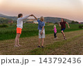 Group of happy children enjoying playful activities in a vast green field with hills and a warm sunset sky in the background, symbolizing family togetherness and joyful childhood moments 143792436