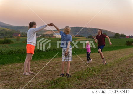 Group of happy children enjoying playful activities in a vast green field with hills and a warm sunset sky in the background, symbolizing family togetherness and joyful childhood moments Group of happy children enjoying playful activities in a vast green field with hills and a warm sunset sky in the background, symbolizing family togetherness and joyful childhood moments 143792436