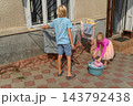 Two young siblings help with household chores on a sunny day, boy hangs clothes on a drying rack while girl lifts laundry from a basin in the yard, learning responsibility together 143792438