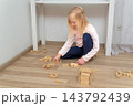 Blonde child concentrating while playing on the light wood floor, building structures with wooden toy blocks, engaging in educational play and development at home 143792439
