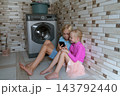 Two young children sitting on the floor in a utility room, sharing a mobile phone screen while a washing machine operates, symbolizing modern childhood and digital entertainment 143792440