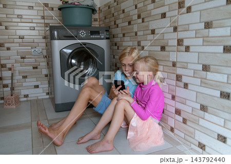 Two young children sitting on the floor in a utility room, sharing a mobile phone screen while a washing machine operates, symbolizing modern childhood and digital entertainment Two young children sitting on the floor in a utility room, sharing a mobile phone screen while a washing machine operates, symbolizing modern childhood and digital entertainment 143792440
