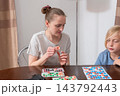 Woman and boy focused on a loto game at a wooden table, placing tokens on numbered cards and sharing a relaxed, playful bonding moment during home leisure time 143792443