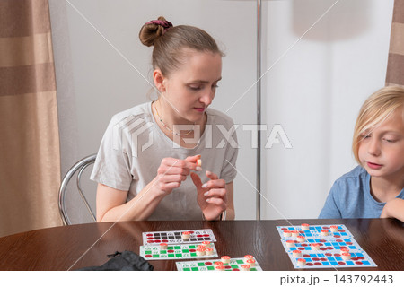 Woman and boy focused on a loto game at a wooden table, placing tokens on numbered cards and sharing a relaxed, playful bonding moment during home leisure time 143792443