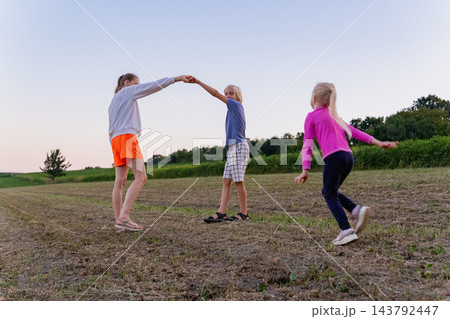 Mother, girl and a boy barefoot and holding hands, dancing and running through a rural field at sunset, captured in warm summer evening light, joyful and carefree 143792447