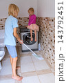 Blond boy mopping the tiled bathroom floor as his blond sister sits on the washing machine, portraying childhood involvement in daily household chores and responsibility 143792451