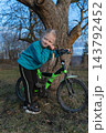 Young girl smiling and leaning on her green bicycle, enjoying an outdoor moment with a large tree and natural background, depicting childhood simplicity and open air fun 143792452