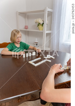 Young boy intently playing dominoes with another person at a wooden table indoors, focused on placing tiles in a quiet family moment of learning, strategy and bonding Young boy intently playing dominoes with another person at a wooden table indoors, focused on placing tiles in a quiet family moment of learning, strategy and bonding 143792453