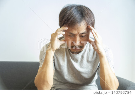 Portrait of 50s mature Asian man headache, pressing on head with painful expression, sitting on sofa at home, medicines and water on table. 143793784