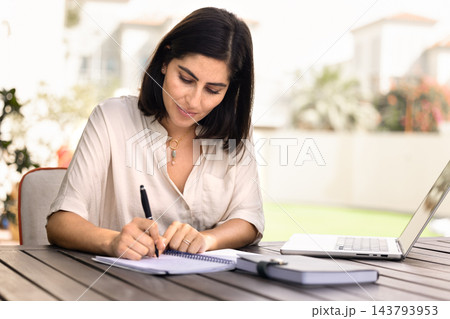 Woman taking notes in notebook sitting at table in courtyard 143793953