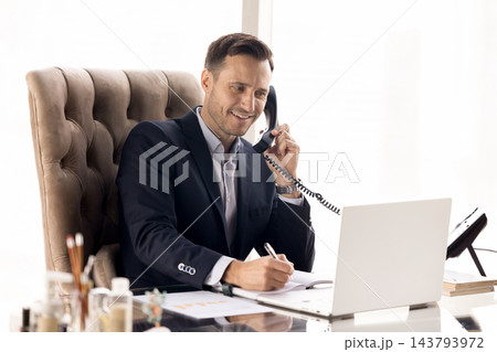 Smiling businessman in suit talking on landline phone at workplace 143793972