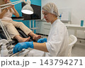 Female podiatrist examines patients feet at clinic, holding small instrument beside gloved hands. Focused foot care professional works under bright lamp in clean treatment room. 143794271
