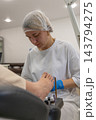 Focused nail technician cleans client toenails during beauty salon pedicure appointment. Aesthetician dressed in white uniform applies skincare cosmetics, showing serious professional concentration. 143794275