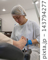 Pedicurist treats client foot carefully during professional beauty procedure. Focused specialist wears medical hairnet and blue gloves while applying cosmetic oil with small wooden brush. 143794277