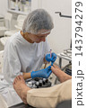 Skilled esthetician performs detailed pedicure procedure on customer feet using specialized precision tool. Dedicated worker carefully tends to toenail hygiene inside bright clinical clinic. 143794279