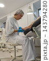 Foot care specialist examines patients foot under bright clinic lamp, holding toes during nail treatment. Attentive podiatrist wears blue gloves and protective cap beside treatment chair. 143794280