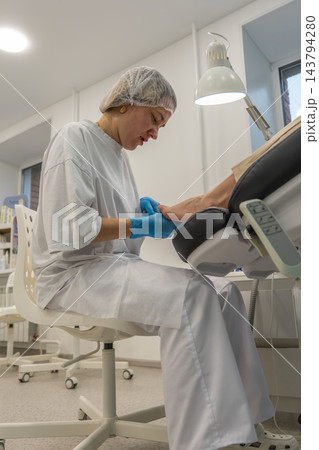 Foot care specialist examines patients foot under bright clinic lamp, holding toes during nail treatment. Attentive podiatrist wears blue gloves and protective cap beside treatment chair. Foot care specialist examines patients foot under bright clinic lamp, holding toes during nail treatment. Attentive podiatrist wears blue gloves and protective cap beside treatment chair. 143794280