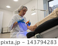 Podiatrist applies blue light to patients toenail while guiding small instrument. Attentive clinician works in clean treatment room with gloves and hair cover, positioning instrument at nail. 143794283