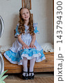 Smiling Girl Seated On Bench Wearing Blue Ruffled Dress, Hands Folded On Lap, Polished Mary Jane Shoes, Soft Curls, Warm Natural Light, Neutral Wallpaper Backdrop, Gentle Confident Expression. 143794300