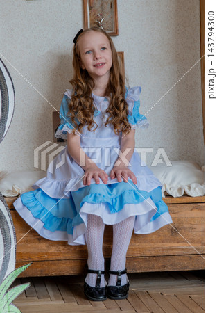 Smiling Girl Seated On Bench Wearing Blue Ruffled Dress, Hands Folded On Lap, Polished Mary Jane Shoes, Soft Curls, Warm Natural Light, Neutral Wallpaper Backdrop, Gentle Confident Expression. 143794300