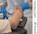 Pedicurist wearing protective gloves treats human foot during clinical pedicure session. Carefully arranged bottles and treatment brushes support thorough toenail health procedure. 143794301